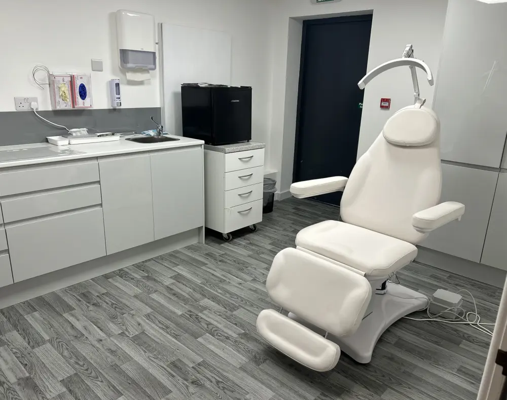 Dental examination chair and medical supplies in a clinical room with white cabinets, a mini fridge, and a door.