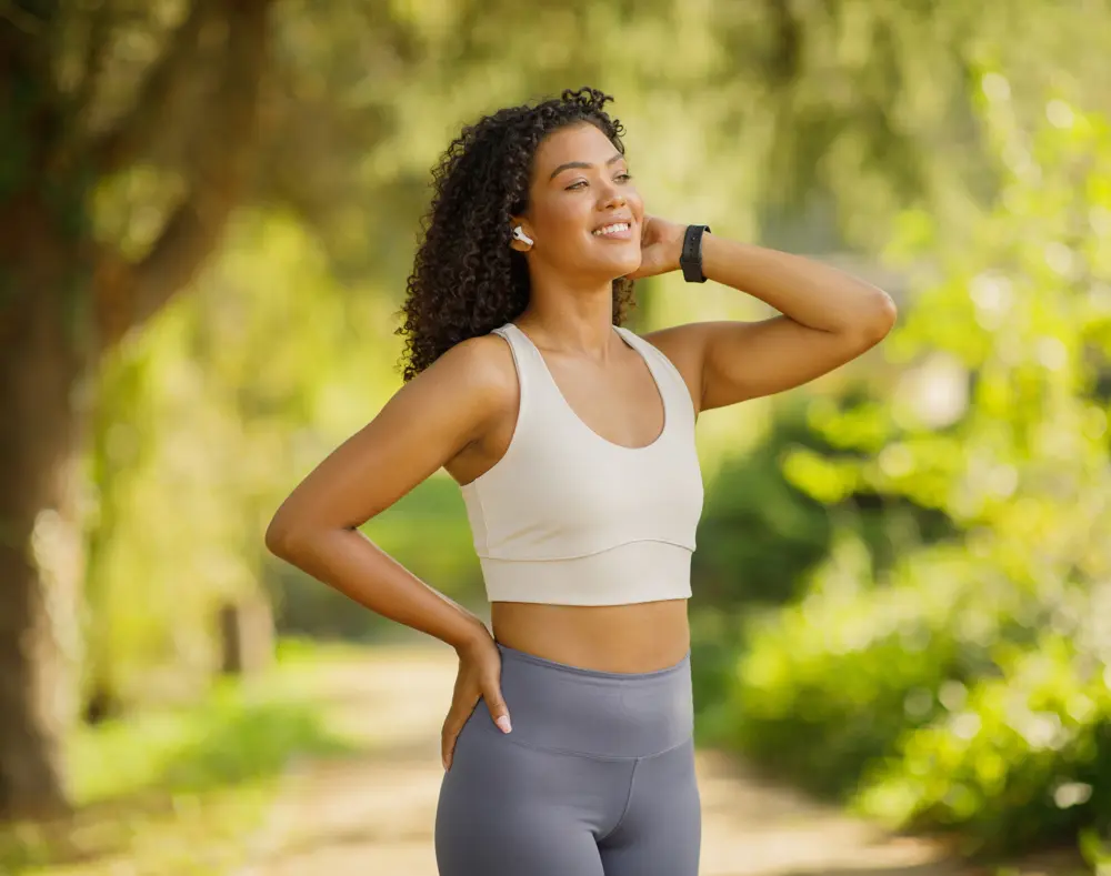 A young woman with curly hair smiling outdoors in a sunny, green park, wearing workout clothes, with one hand on her hip and the other touching her neck.