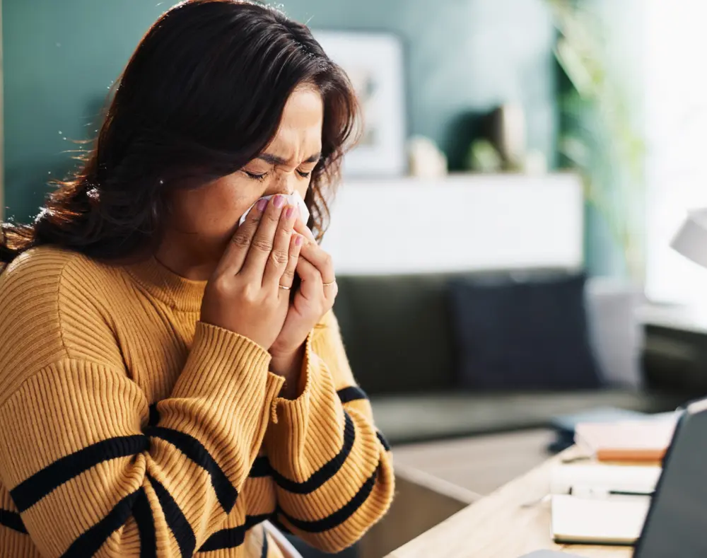 Woman in a mustard striped sweater holding a tissue to her nose, appearing to be sneezing or experiencing allergy symptoms indoors.
