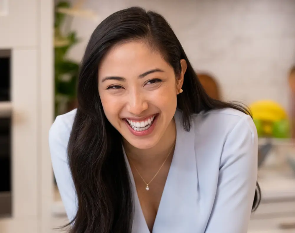 A woman with long dark hair, smiling and looking slightly to the side, in an indoor setting with blurred background elements.