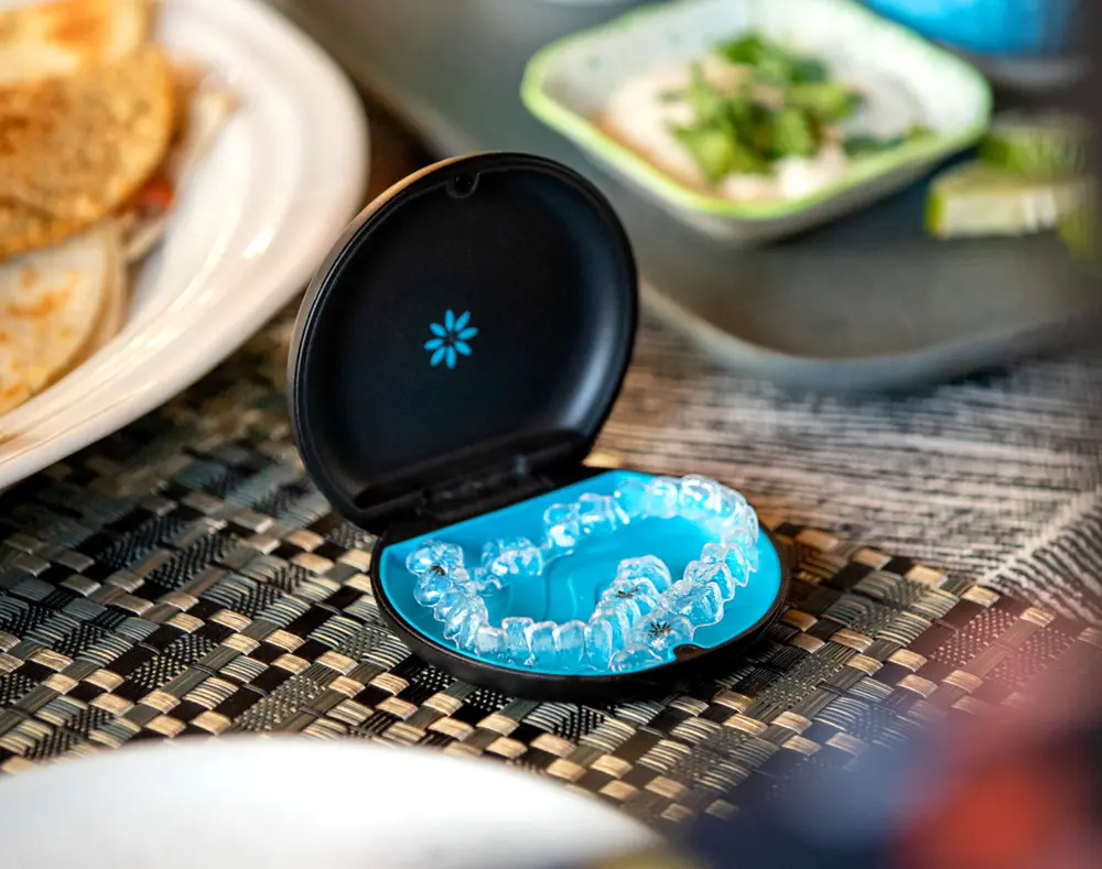 Clear dental aligners in a black case with a blue interior, placed on a woven table mat with dishes of food in the background.