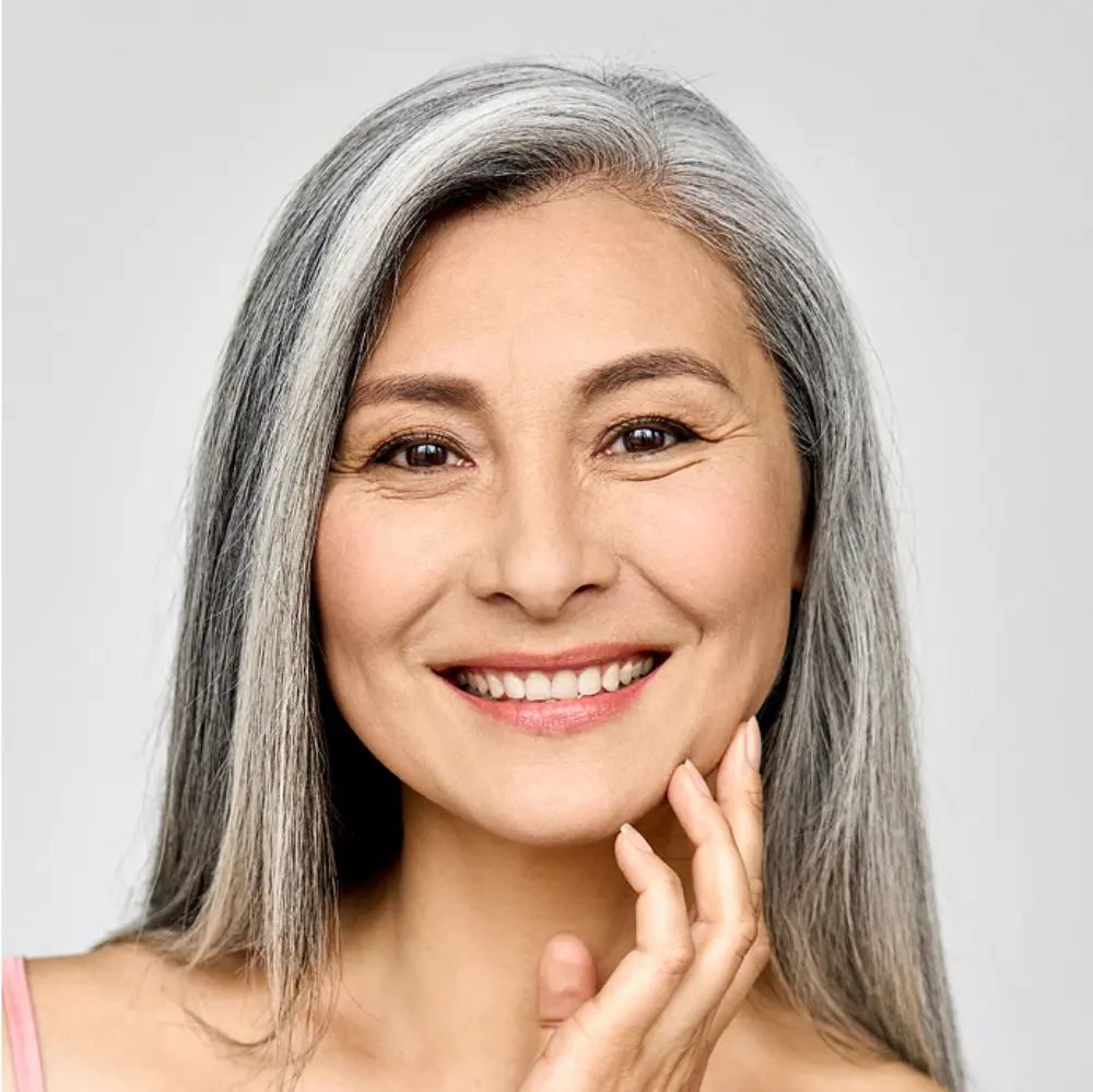 A close-up of a smiling mature woman with long gray hair, touching her face gently, against a plain light background.