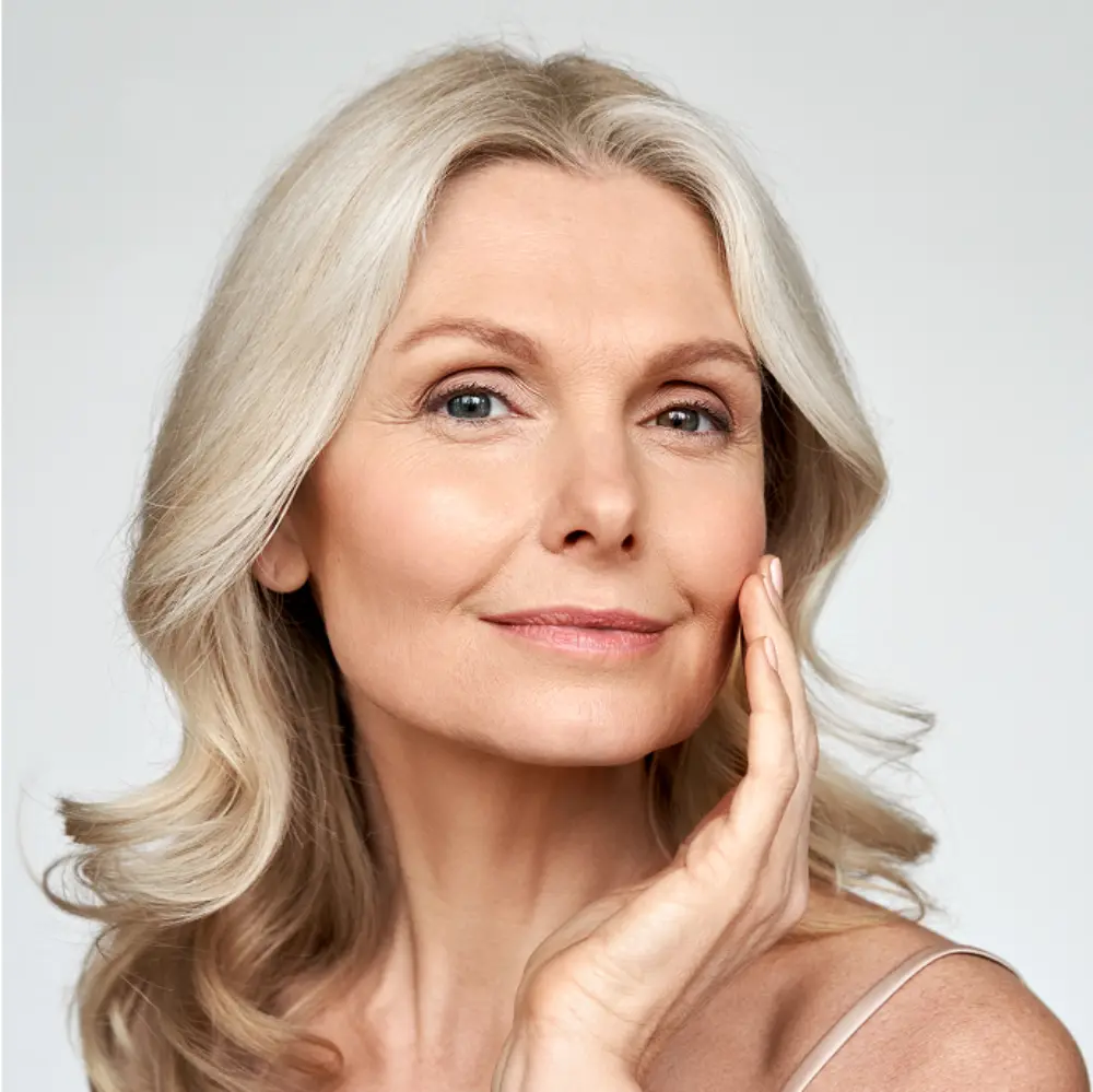 A mature woman with long, wavy gray hair gently touches her face, smiling softly against a plain light background.