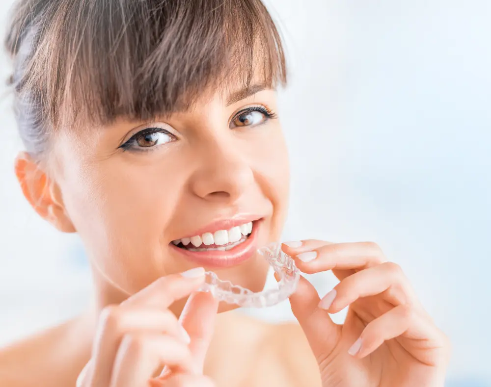 Woman smiling and holding a clear dental aligner near her mouth against a bright background.