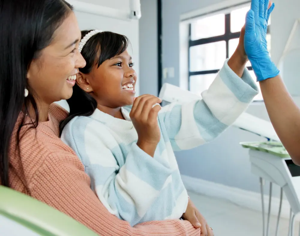 A young girl, wearing a white sweater, happily raises her hand with a gloved hand while smiling, sitting close to a smiling woman in a medical setting.