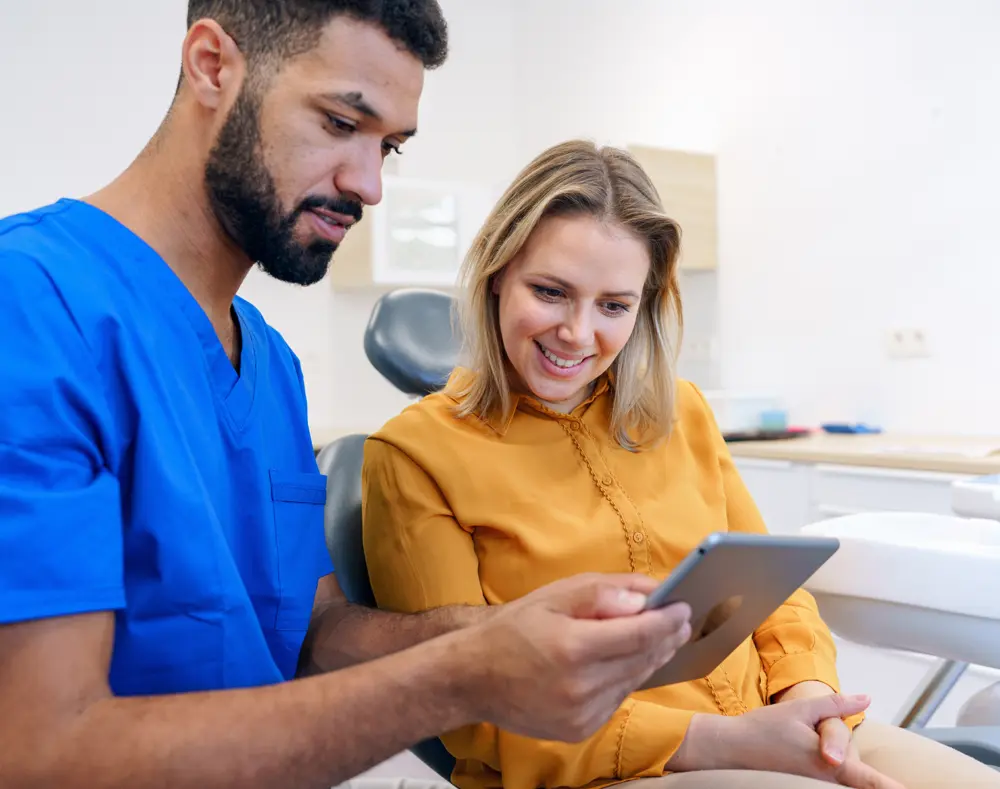 A healthcare professional and a woman sit together in a medical setting, examining a tablet with smiles, surrounded by medical equipment and white walls.