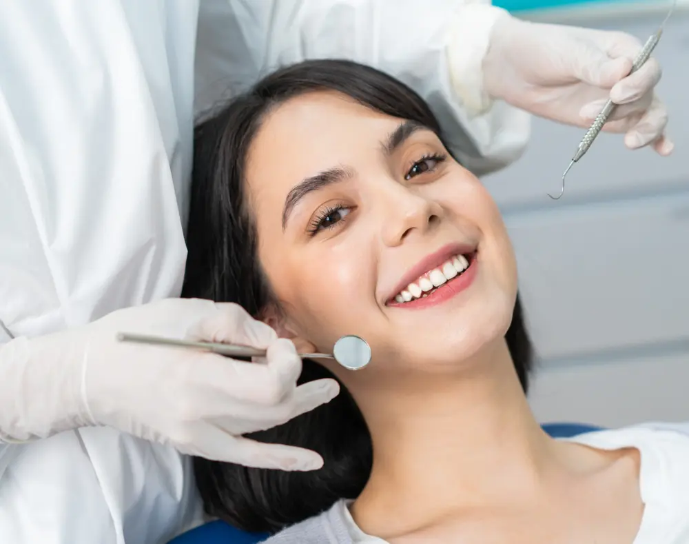 Smiling woman in dental chair during checkup, with dentist holding dental tools near her face in a clinical setting.