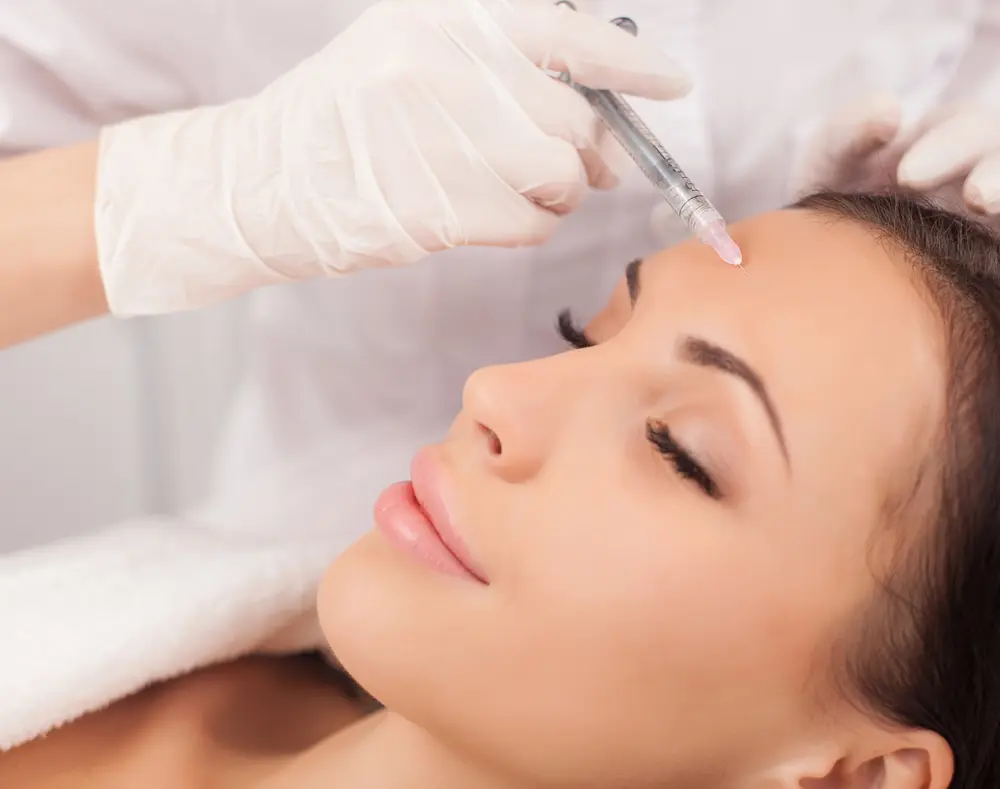 A woman receives a facial injection treatment from a medical professional wearing gloves, lying back with eyes closed, in a clinical setting.