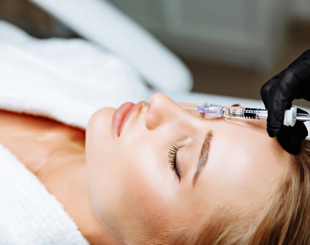 Woman receiving a facial injection on her forehead in a spa or medical setting, with her eyes closed and a gloved hand administering the treatment.