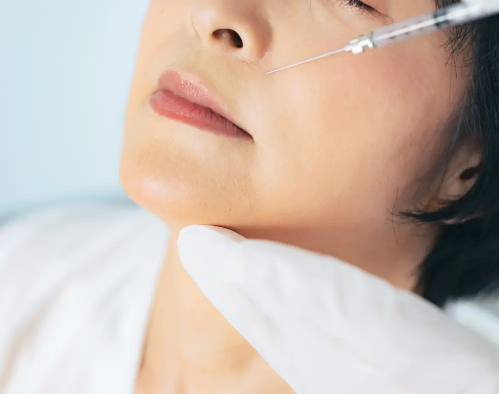 A close-up of a woman receiving a facial injection, with a healthcare professional's gloved hand supporting her chin and the needle near her cheek.