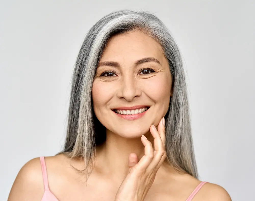 A smiling mature woman with long gray hair, touching her face with her hand, wearing a pink spaghetti strap top against a plain light background.