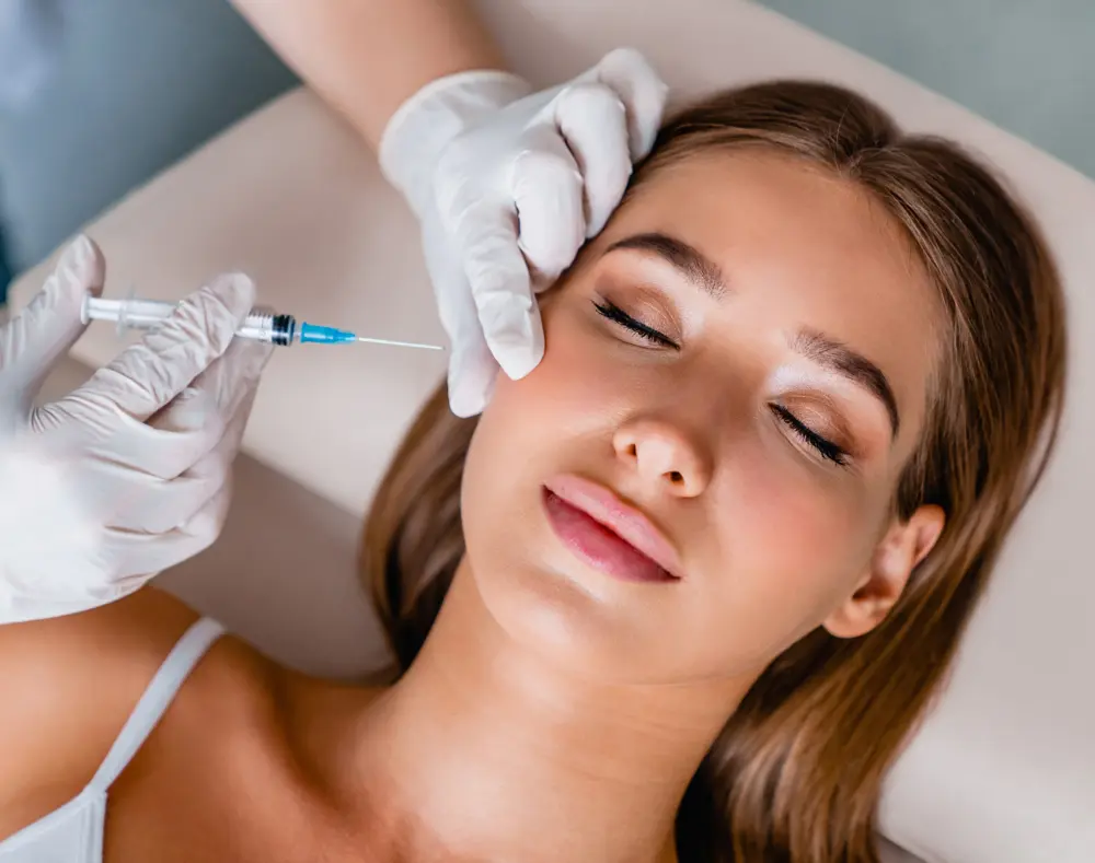 Woman receiving a facial treatment with a syringe, lying on a bed, eyes closed, with a medical professional wearing gloves performing the procedure.
