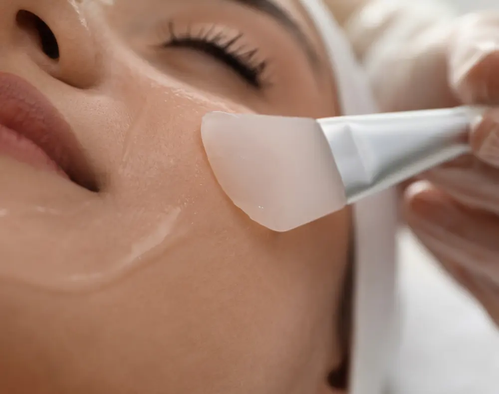 A person receiving a facial treatment with a spatula applying a skincare mask onto their cheek. The focus is on the person's face and the applicator.