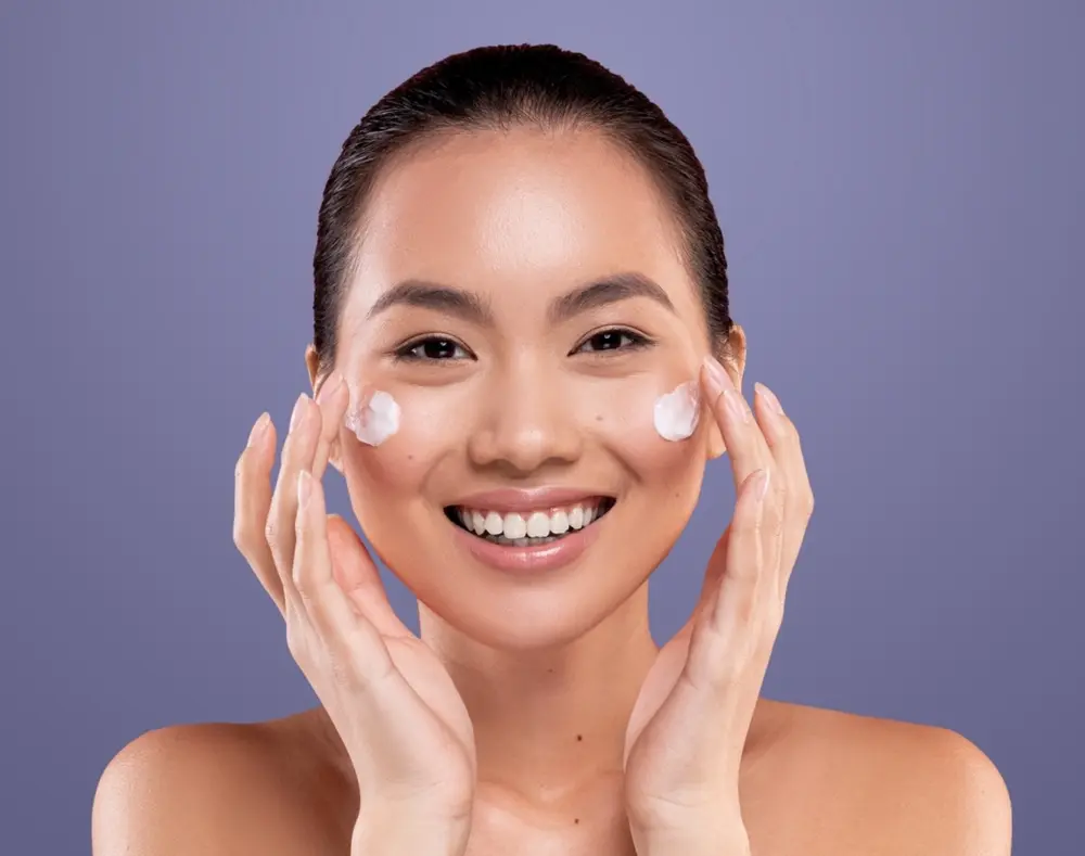 A smiling young woman with dark hair, applying cream to her cheeks against a purple background.