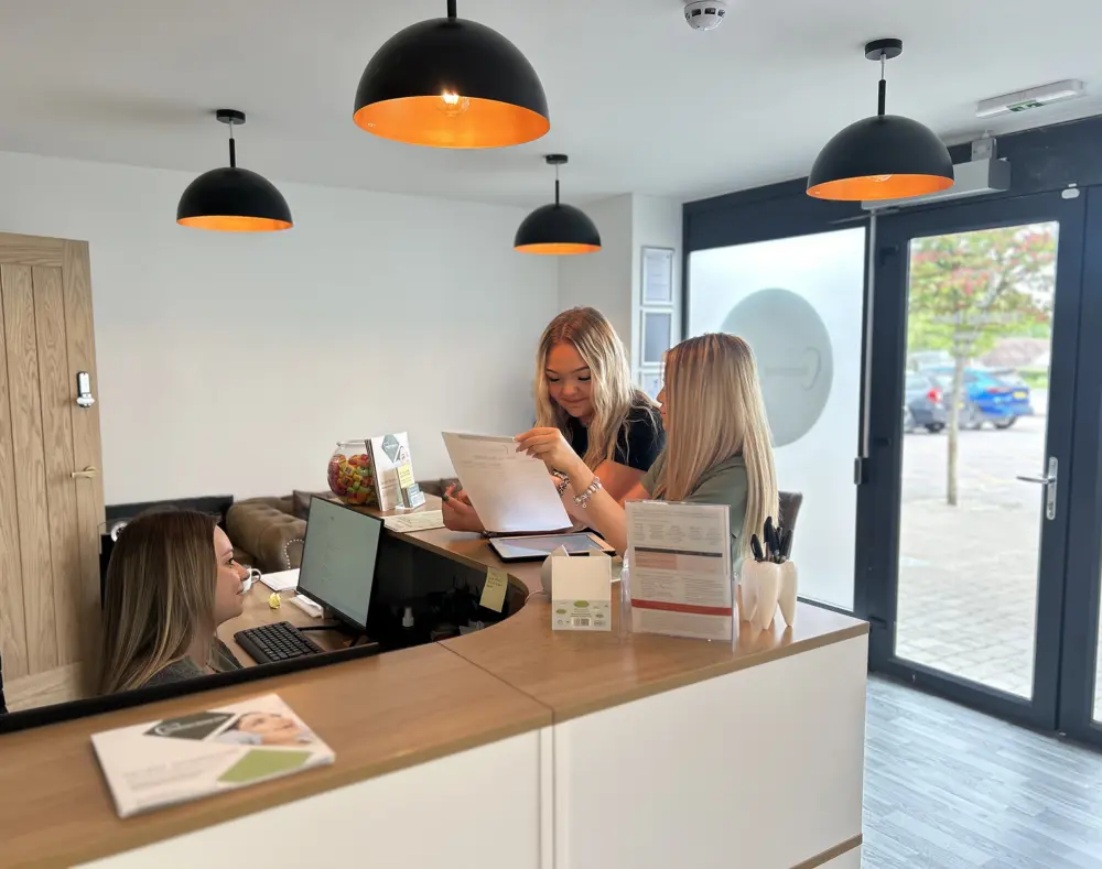 Three women at a reception desk inside a modern office or clinic, engaging with documents and computers near a large glass door with outdoor parking visible.
