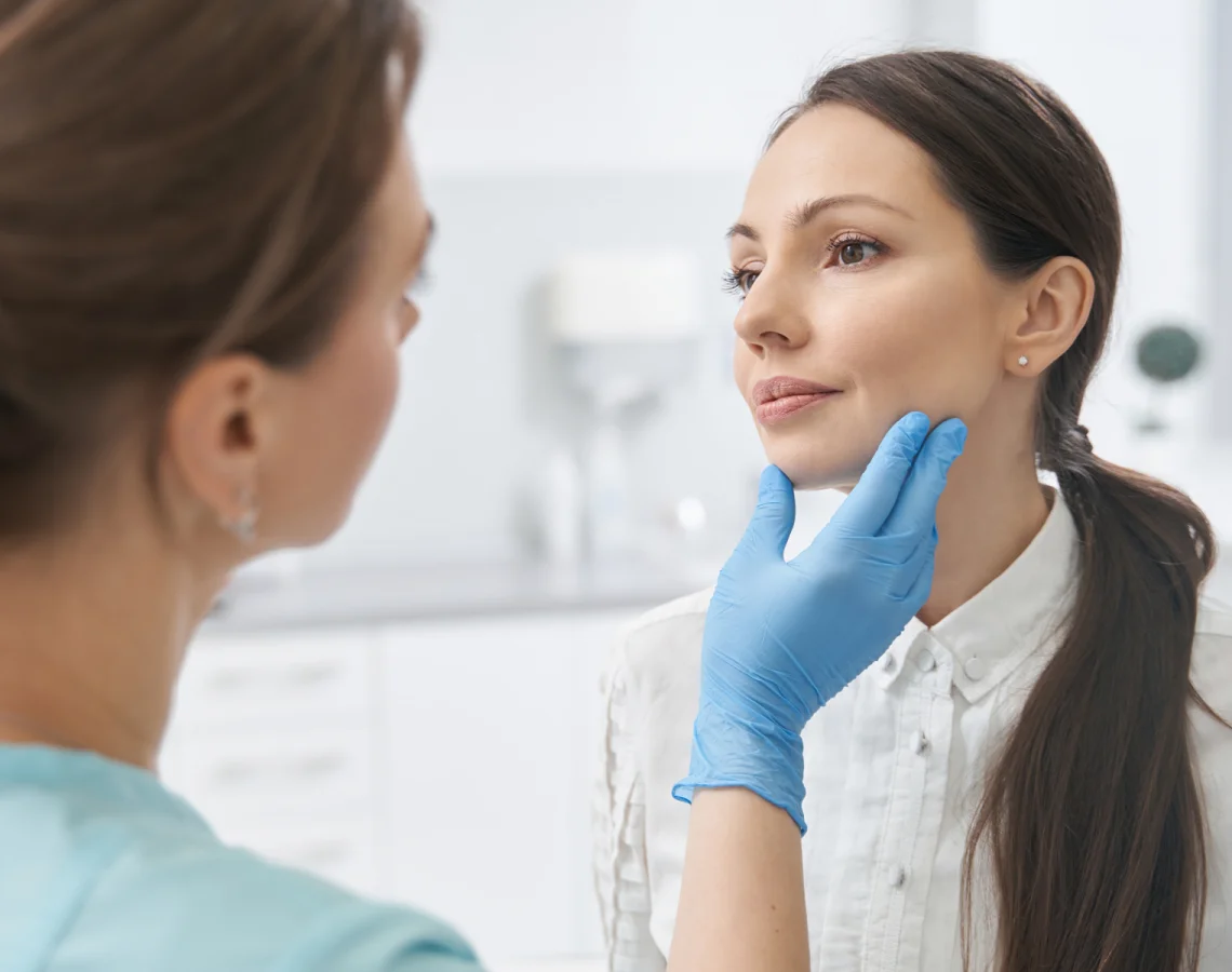 A healthcare professional examines a woman’s face, gently holding her chin with gloved hands in a clinical setting with white background.