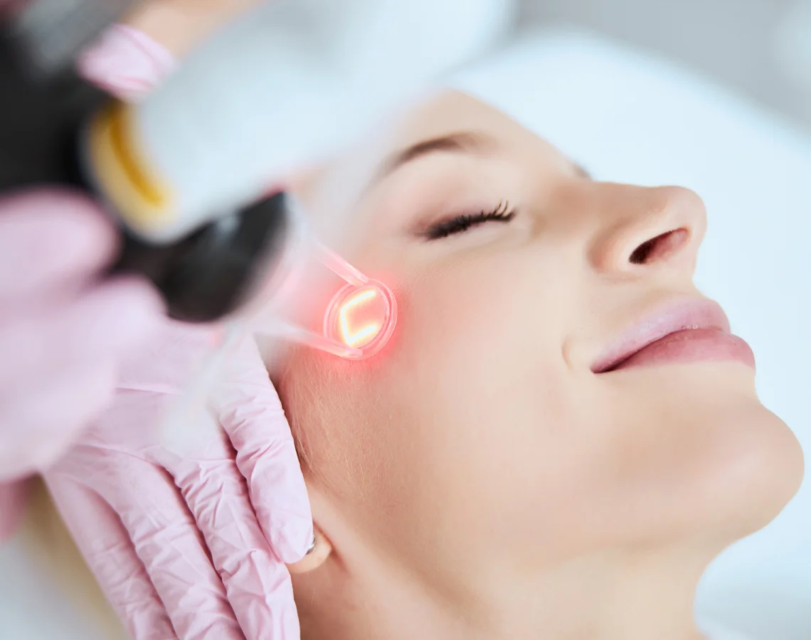 A woman receiving laser skin treatment on her cheek, lying with eyes closed, as a medical professional uses a laser device with a red light.