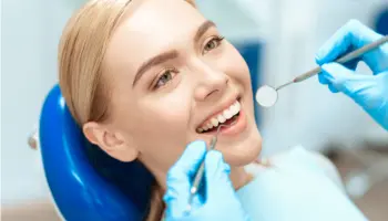 Woman at dentist's office receiving dental check-up, lying in a dental chair, smiling while a dentist examines her teeth with dental tools.