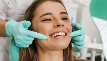 A woman smiling during a dental check-up, with a dentist using gloved hands to examine her teeth in a bright dental office.