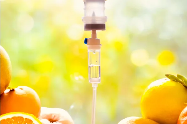 An IV drip hangs over a variety of fresh oranges and lemons, with a blurred outdoor background, suggesting a health or nutrition theme involving fruit.