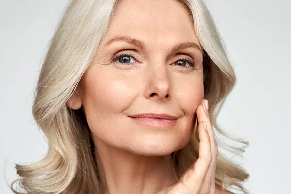 A mature woman with long, wavy gray hair gently touching her face, smiling softly, against a plain light background.