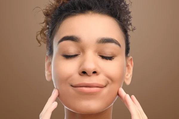 A woman with curly hair, eyes closed, smiling softly, gently touching her face with both hands, peaceful expression against a neutral background.