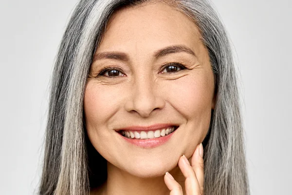 A smiling mature woman with long gray hair, gently touching her face, wearing a pink top, against a plain white background.