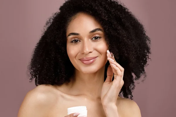 A smiling woman with curly hair applying moisturizer to her face against a muted pink background.