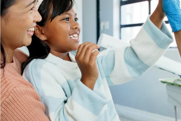 A young girl in a white sweater smiling as a healthcare worker in blue gloves prepares her for a dental or medical procedure with an assistant.