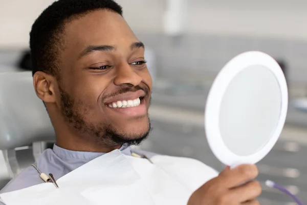 Smiling man in a dental chair holding a mirror, looking at his reflection with a happy expression, in a dental clinic setting.
