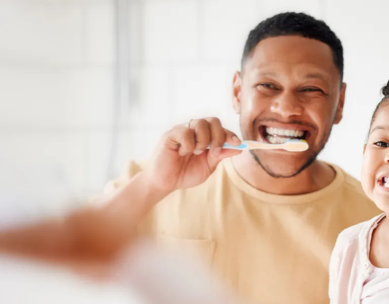 A man and young girl brush their teeth together, smiling and enjoying the activity in a bright bathroom setting.